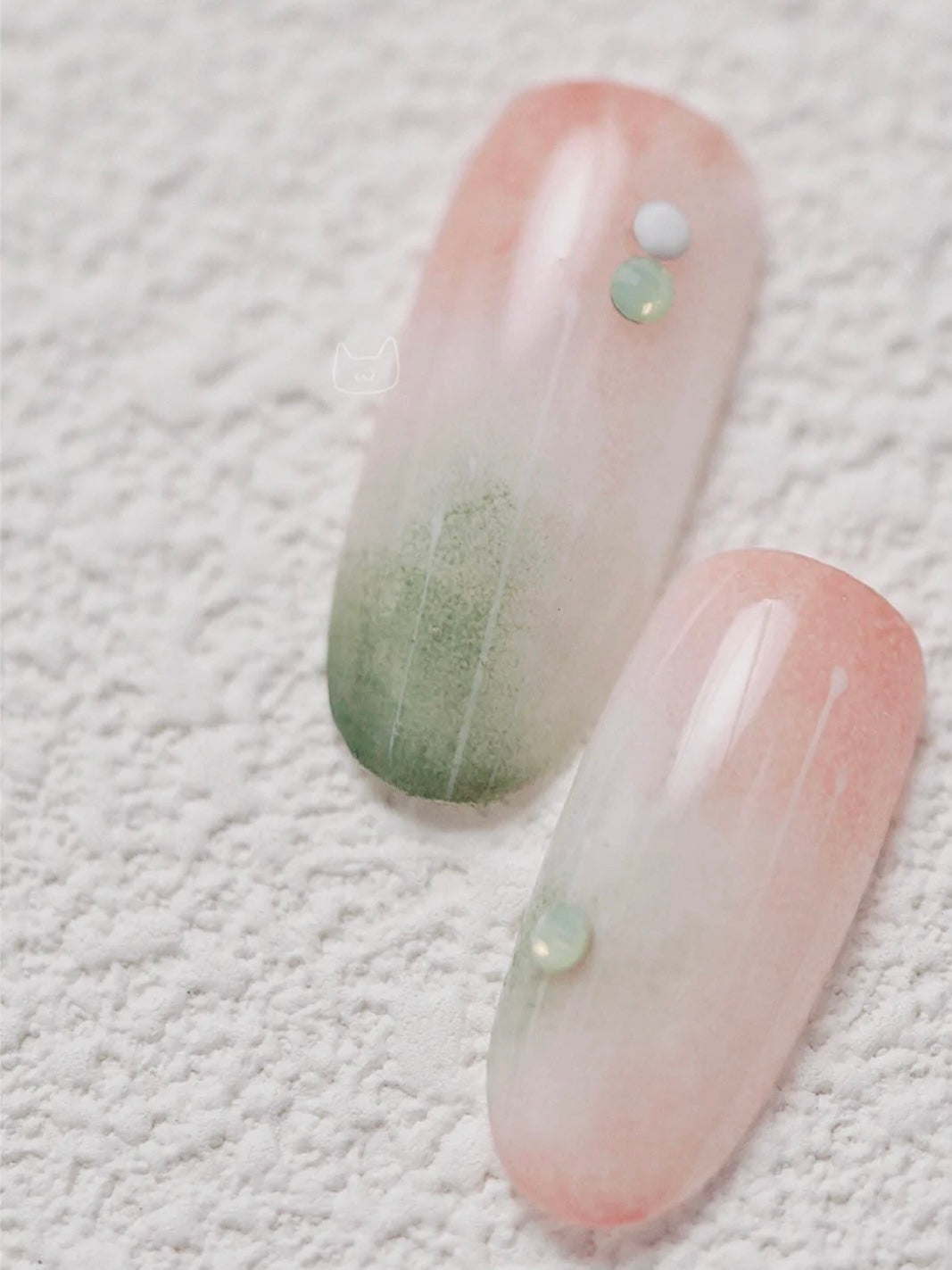 Two elongated nail art samples resting on a textured white surface, showcasing gradient designs; one fades from a pale green to a soft peach, and the other from a subtle pink to a muted green, both adorned with a tiny, glossy embellishment near the tip.