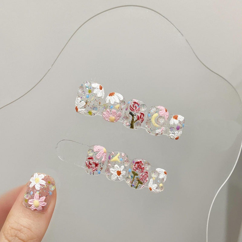 Close-up view of a hand modeling 'Eden' press-on nails, featuring a transparent base adorned with intricate floral designs and colorful gem embellishments, reflecting a garden-like aesthetic.