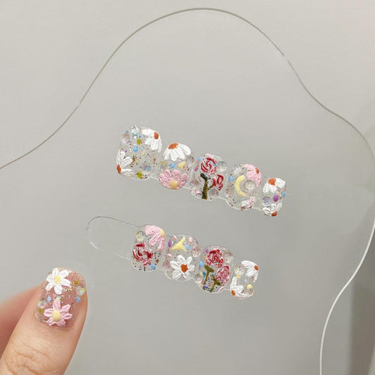 Close-up view of a hand modeling 'Eden' press-on nails, featuring a transparent base adorned with intricate floral designs and colorful gem embellishments, reflecting a garden-like aesthetic.