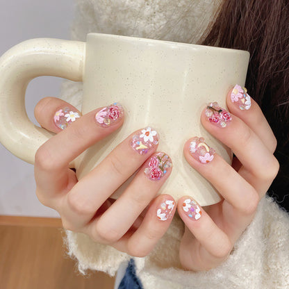 Woman holding a cream mug, showcasing her delicate 'Eden' press-on nails decorated with pink flowers and shiny crystals on a clear base, perfect for a fresh and elegant look.