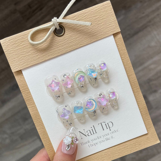 Elegant 'LUNA CHARMS' press-on nails displayed on a card, featuring translucent tips with sparkling moon and star charms, ideal for a whimsical and stylish look.