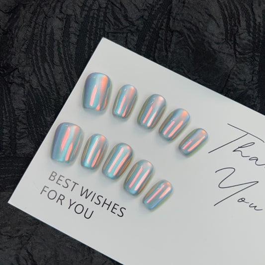 Set of angel chrome press-on nails displayed on a card with 'Best Wishes For You' written on it, featuring a unique iridescent finish that reflects light with hints of pastel colors. These ready-to-apply nails offer a quick and stylish manicure option, laid out elegantly on a textured black lace background.