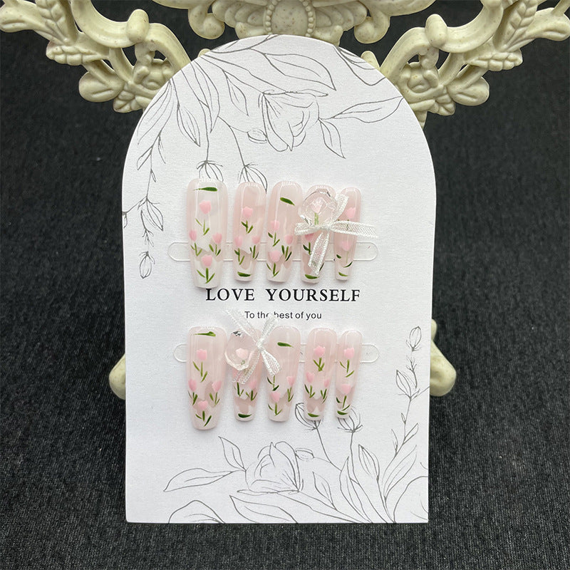 Stylish set of pink 'Flora' press-on nails adorned with subtle white flowers and greenery, presented on a beautifully illustrated card, ideal for a gentle and romantic look