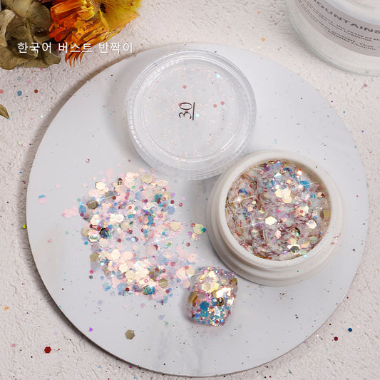 A small container of iridescent nail glitter, featuring a mix of hexagonal and round shapes in various sizes. The glitter is a combination of pastel colors, including pink, blue, and gold, creating a sparkling and eye-catching effect. The container has a clear lid labeled "30" and is shown with some glitter spilled out onto a white surface, showcasing its dazzling and multifaceted appearance. This glitter is perfect for adding a touch of shimmer and glam to any nail design.