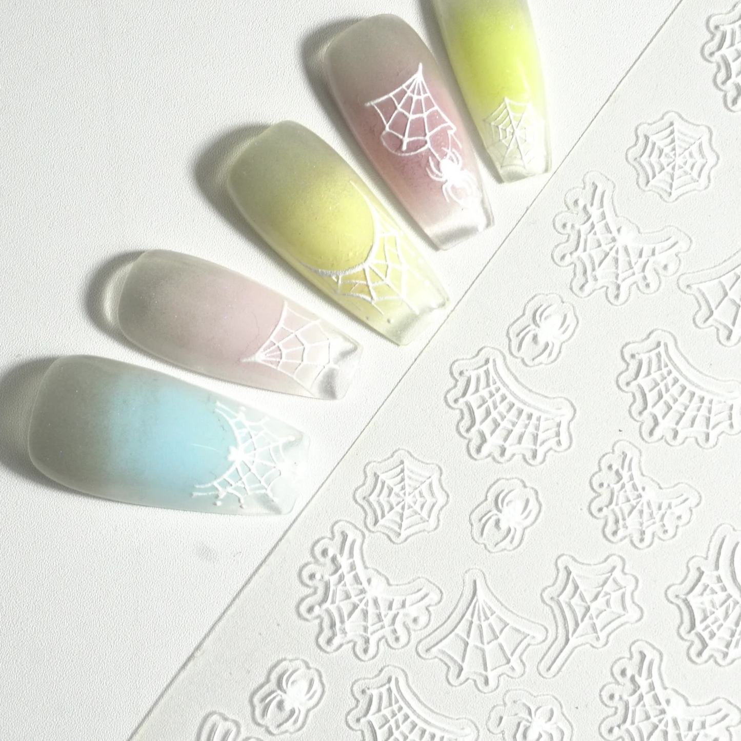 Close-up of white embossed spider web nail stickers with pastel nail examples